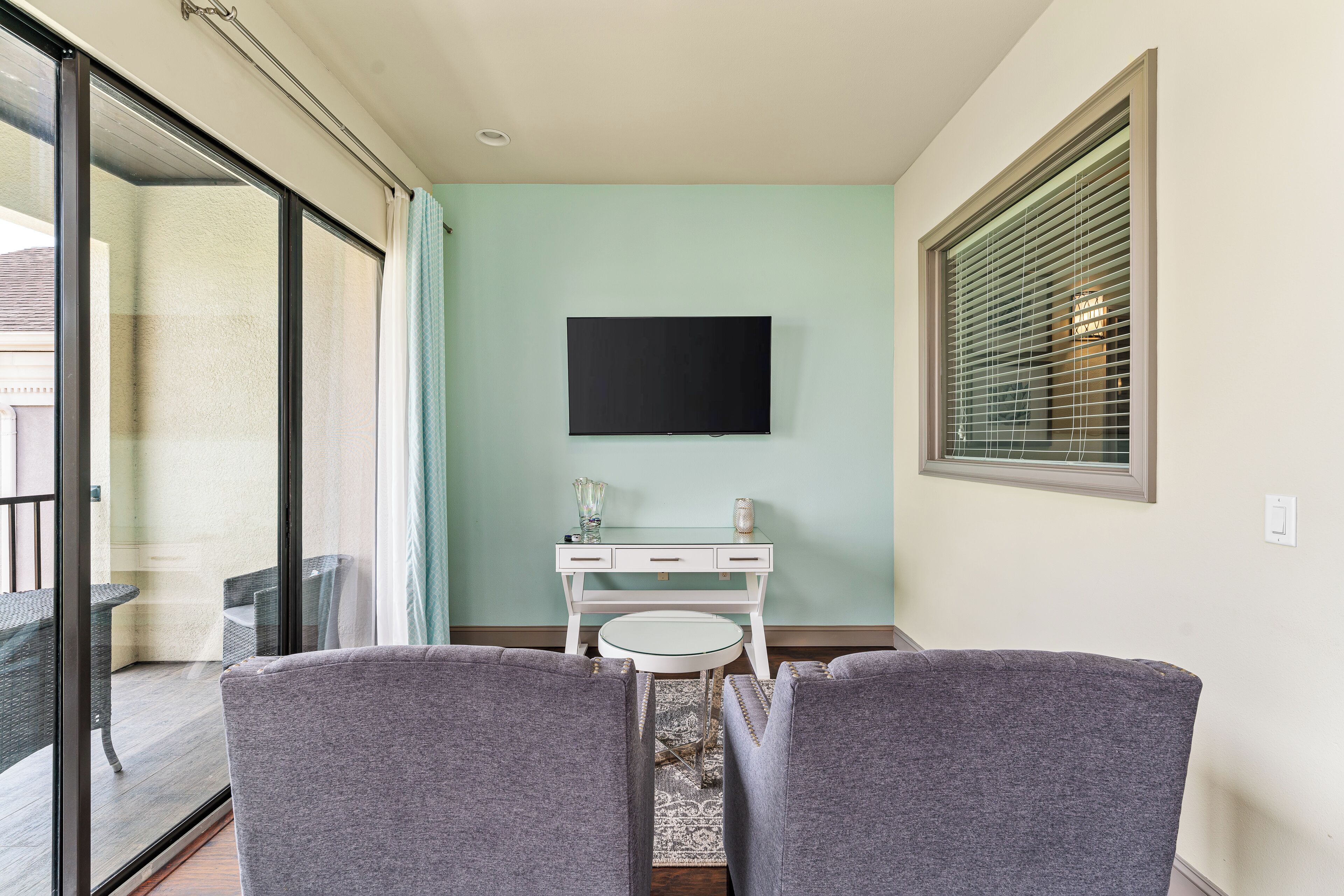 Ein Zimmer mit einer Schiebeglastür, die auf einen Balkon führt, einem weißen Schreibtisch mit einem flachbildfernseher darüber und zwei grauen Sesseln, die zum Schreibtisch hin ausgerichtet sind.