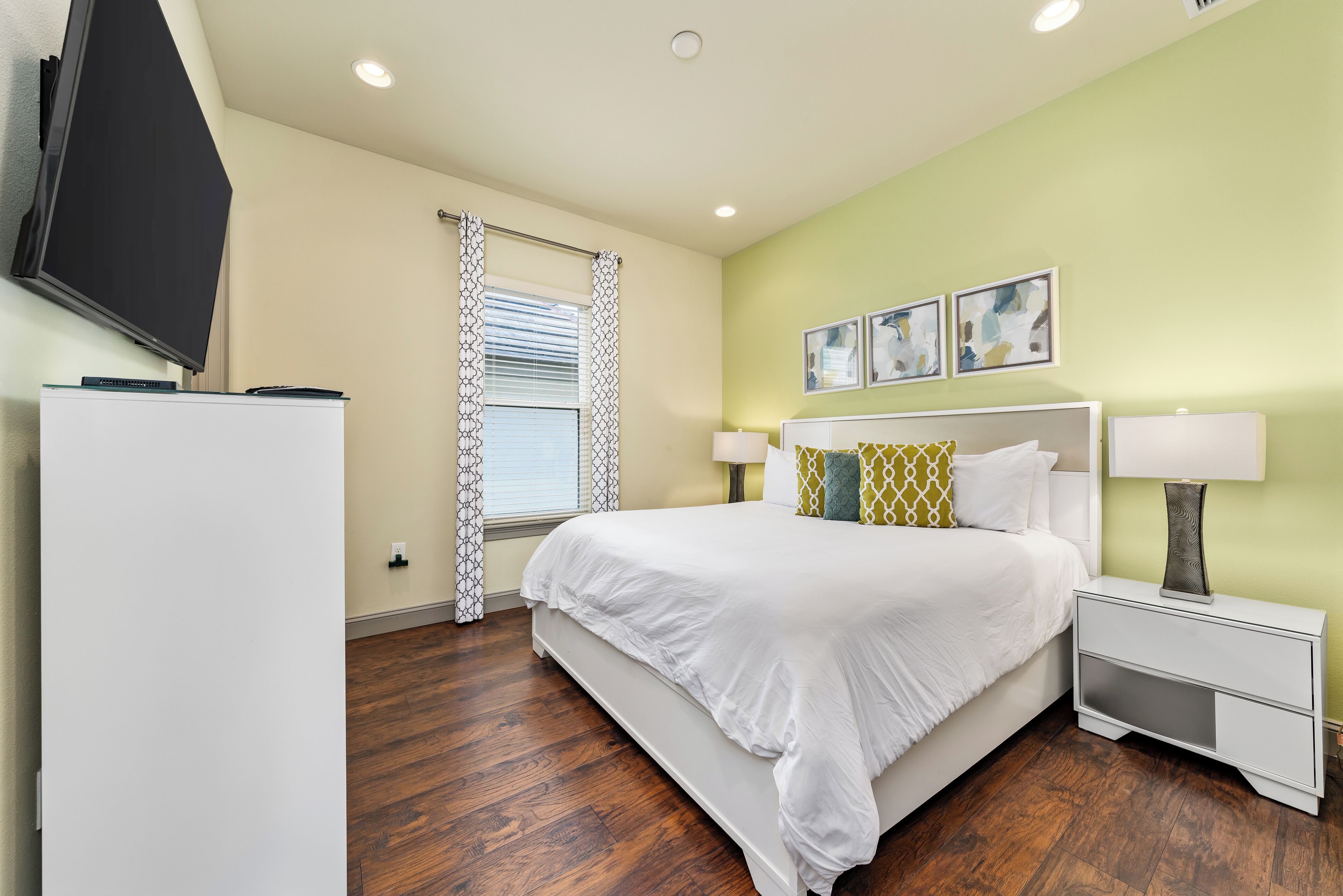 A bedroom with a large bed, bedside tables with lamps, a flat-screen TV mounted on the wall, and artwork above the bed.