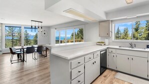 Eine moderne Küche mit weißen Schränken, ein Essbereich mit Tisch und Stühlen sowie große Fenster mit Blick auf Bäume und Berge.