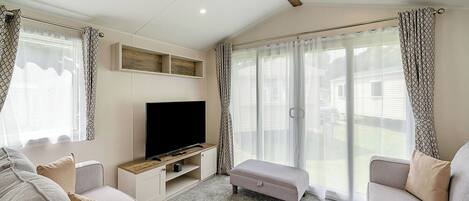 Una sala de estar con una televisión de pantalla plana, dos sofás, un banco y ventanas amplias con cortinas.