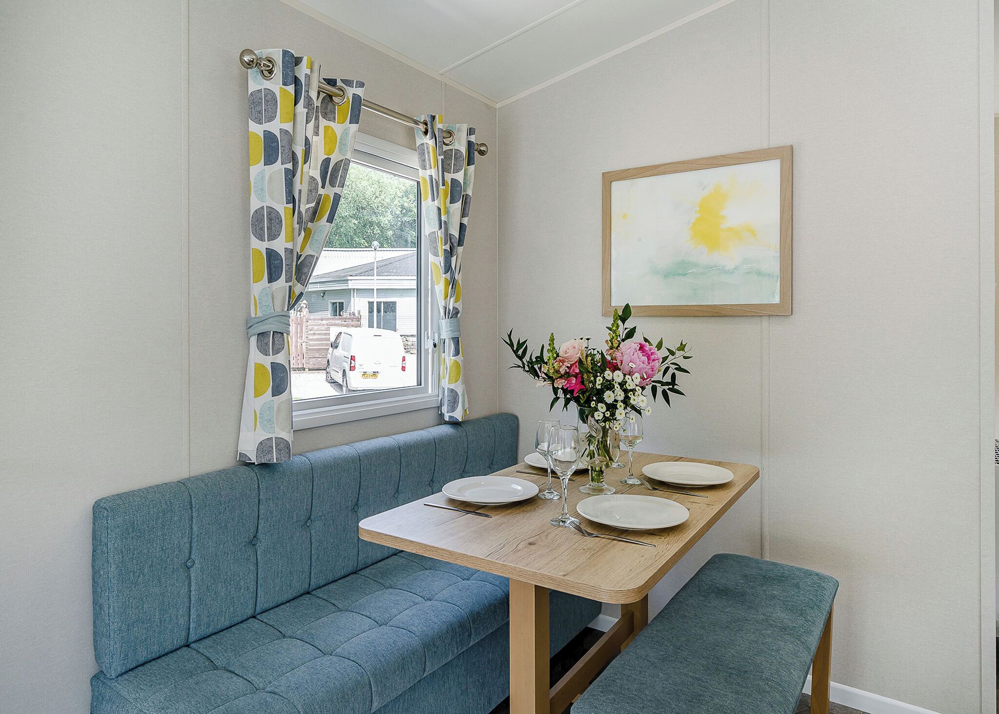 A dining area with a wooden table, blue bench, and a window with patterned curtains.