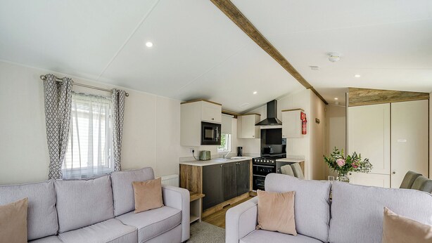 A modern living room with two grey sofas, a kitchen area with a stove and oven, and a wooden ceiling beam.