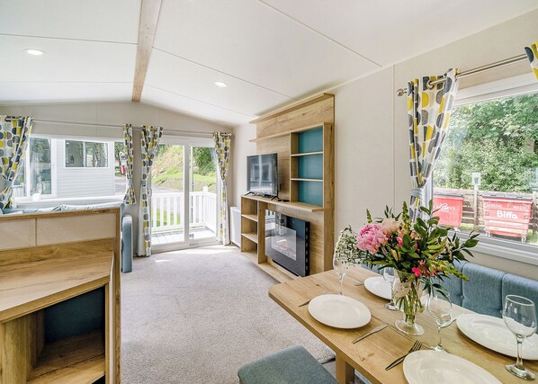 Una habitación acogedora con televisor, estantes de madera, una mesa de comedor lista para dos personas y una ventana con cortinas.