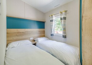 A small, single-room accommodation with two beds, a wooden headboard, a window with striped curtains, and a teal accent wall. - 2 Bedroom platinum unit with decking. (Longridge, Ribble Valley)