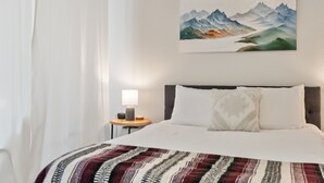 Une chambre d’hôtel avec un lit, un tableau représentant des montagnes accroché au mur, une table de chevet avec une lampe et une fenêtre avec des rideaux.