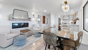 A modern living room with a large TV, comfortable seating, a dining area with wicker chairs, and a kitchen in the background. - "You are My Sunshine" Fully Renovated, 3 Bedroom, 2 Bathroom, Oceanfront Home (Holden Beach)