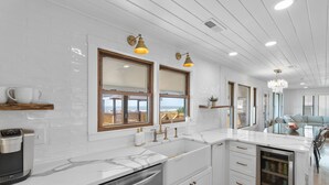 A modern kitchen with white cabinetry, a marble countertop, and a built-in dishwasher. There are two windows with wooden frames and brass fixtures, and a chandelier hanging from the ceiling. - "You are My Sunshine" Fully Renovated, 3 Bedroom, 2 Bathroom, Oceanfront Home (Holden Beach)