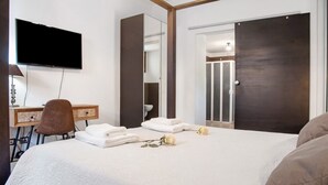 Quarto de hotel com cama, escrivaninha, cadeira, televisão, guarda-roupa e chuveiro.
