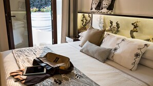 A hotel room with a bed, pillows, a bedside table, a lamp, and a view of the outdoors.