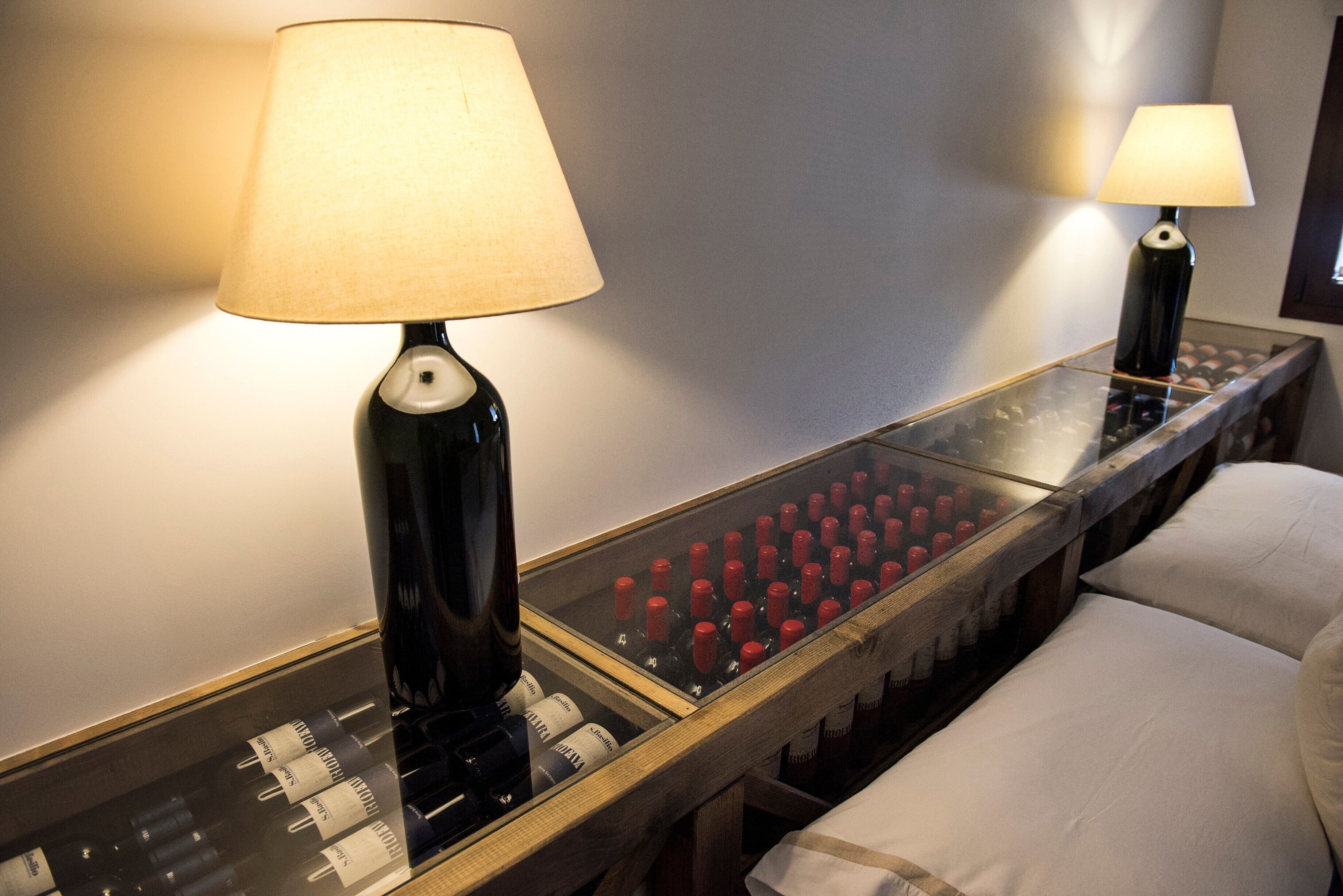 A hotel room with a glass-topped side table displaying bottles, a lamp, and a mirror reflecting the room.