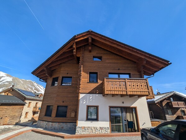 A two-story wooden building with a balcony, stone foundation, and a mountain backdrop.