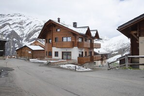 A multi-story wooden chalet with balconies, a stone staircase, and a view of snow-covered mountains.