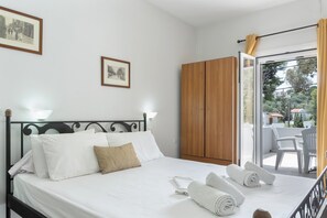 A neatly made bed with white linens and a brown pillow, a wooden wardrobe, and a balcony with white chairs.