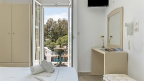 Habitación de hotel con cama, escritorio, espejo, televisor y balcón con vistas a árboles y edificios.