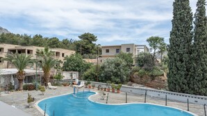 A swimming pool with a curved design, surrounded by potted plants and palm trees, with buildings in the background. - Apartment 'No 12' with Shared Pool, Wi-Fi and Air Conditioning (Kalymnos)
