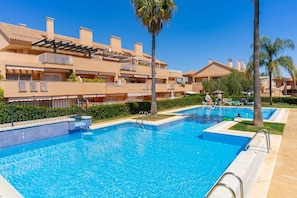 A swimming pool area with multiple pools, a covered seating area, and a building with balconies.