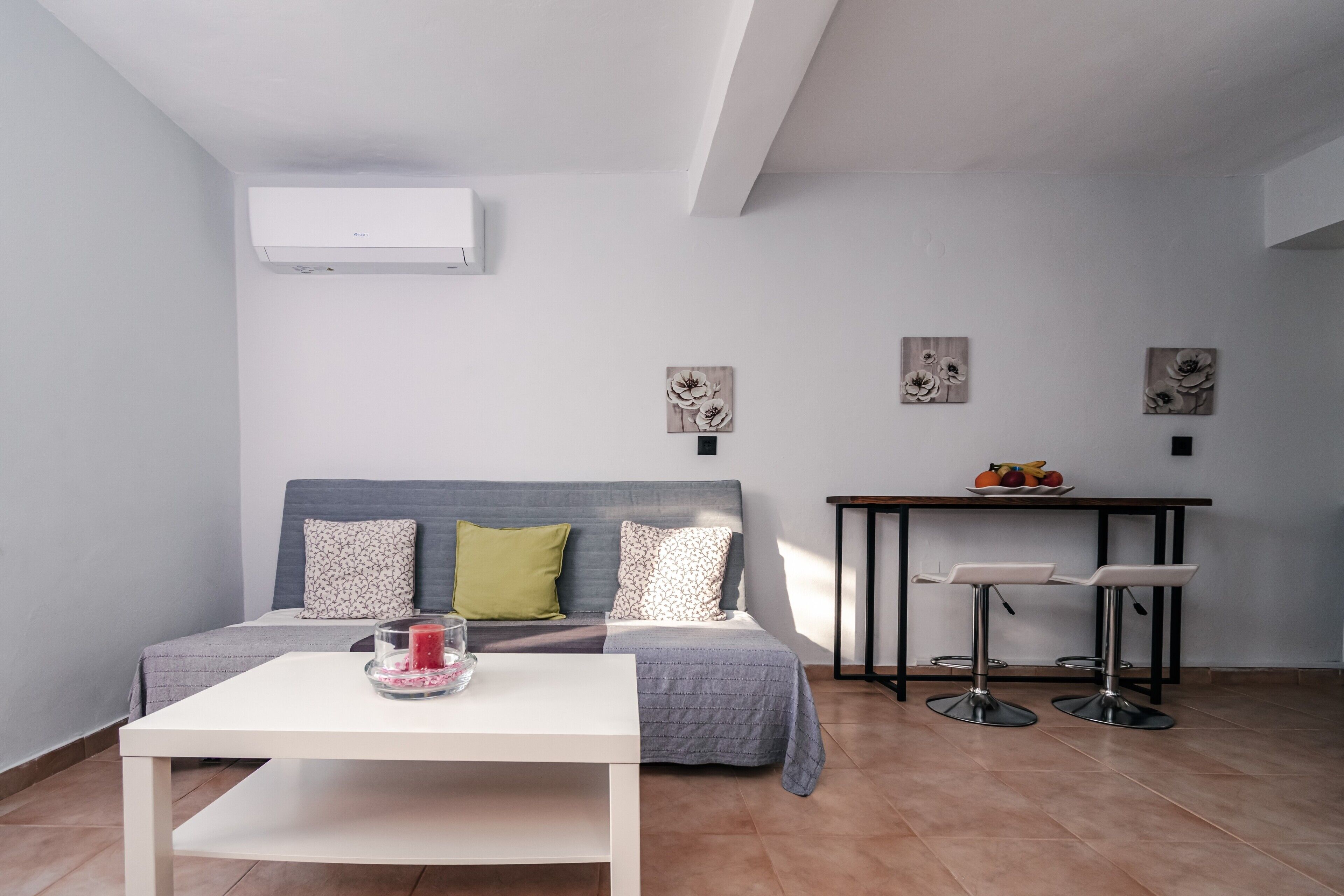 A modern living room with a sofa, a small white table, a dining area with a table and two bar stools, and an air conditioning unit on the wall.