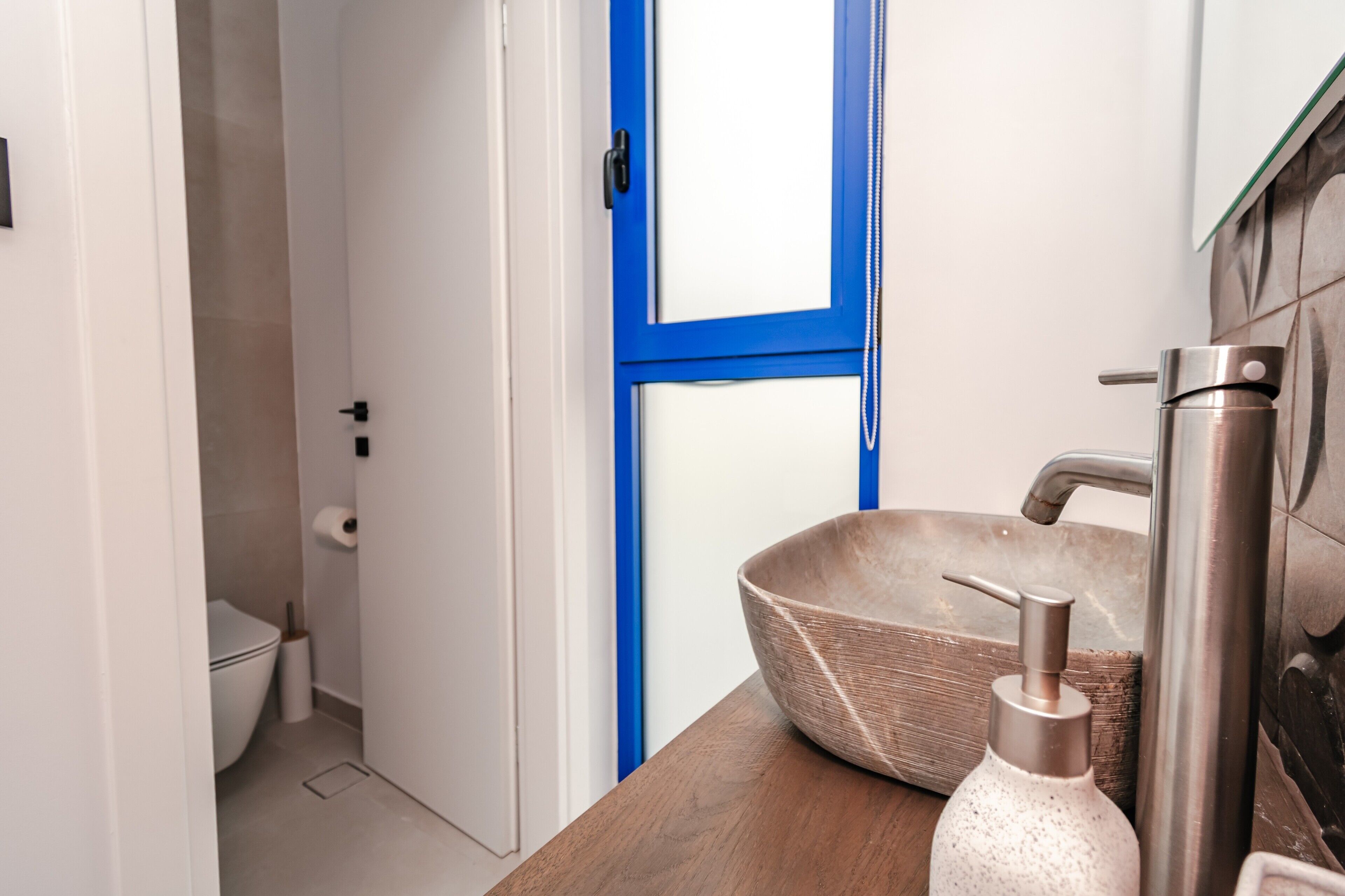 A bathroom with a white toilet, a blue-framed window, a wooden vanity, a large woven bowl sink, and a stainless steel faucet.