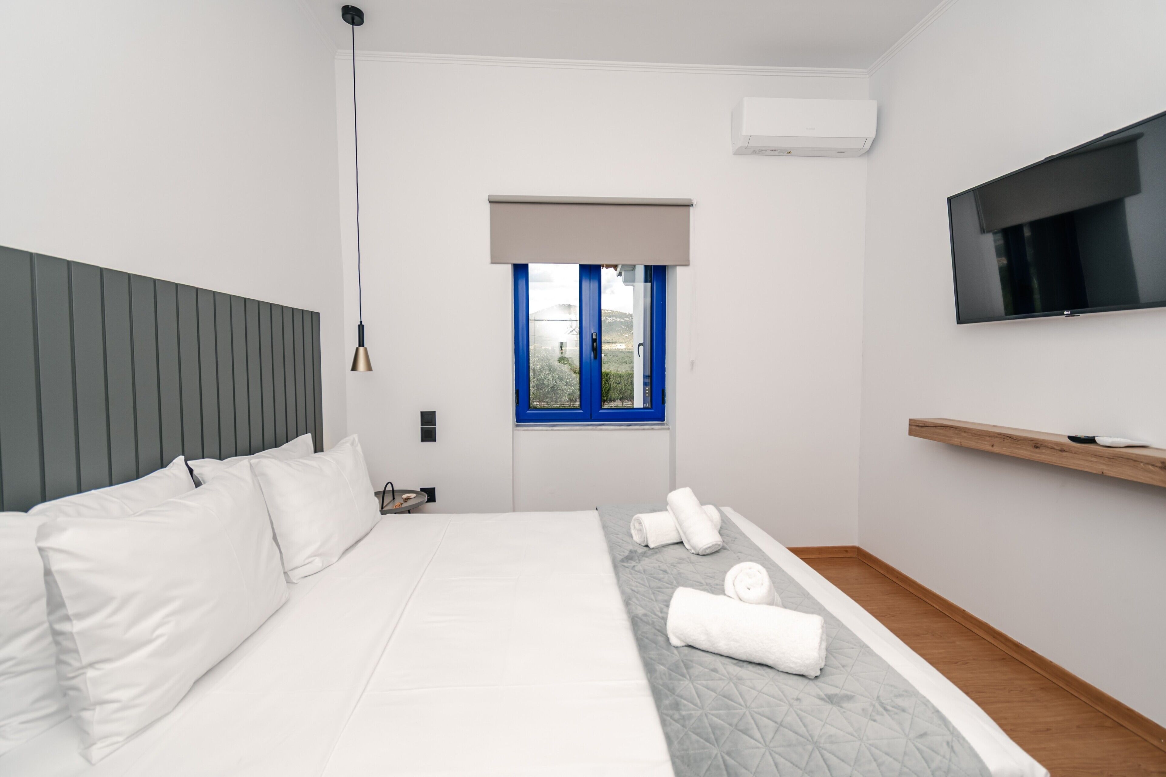 Una camera d'albergo moderna con un letto, una TV, una finestra con persiane blu e una mensola in legno.