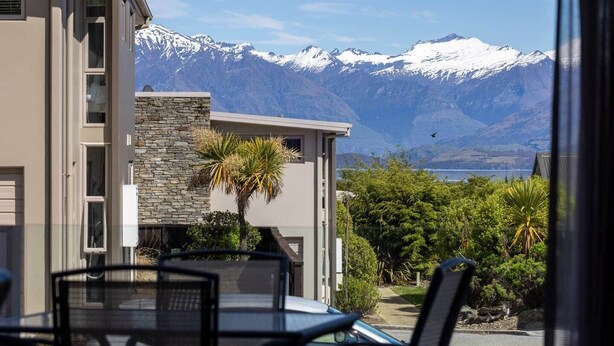 A modern building with a stone facade, a balcony with a table and chairs, and a view of snow-capped mountains.