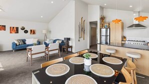 A modern kitchen with a central island, a dining area with a black table and woven placemats, and a living area with a blue sofa and a lamp.