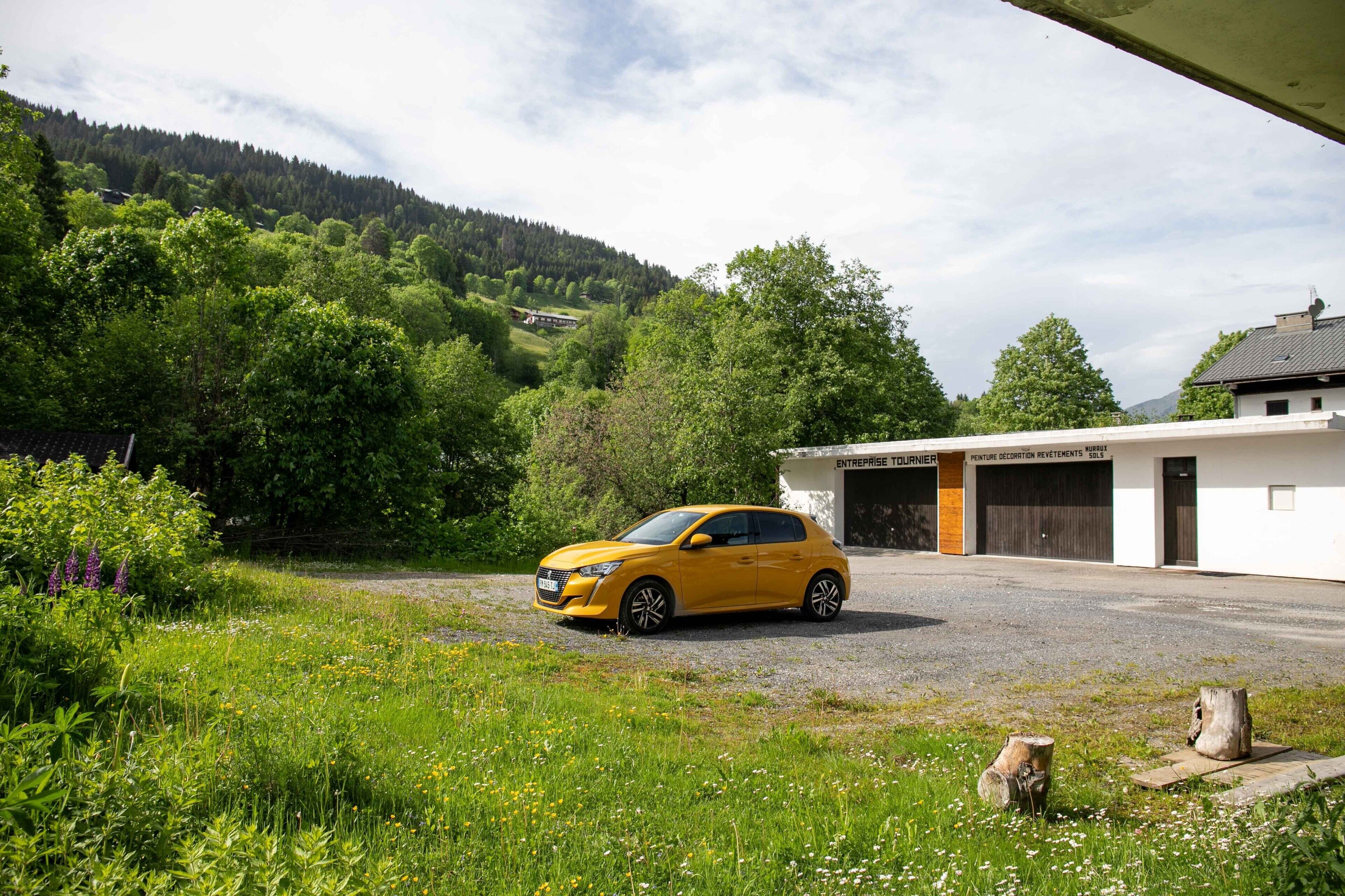 A yellow car parked in front of a building with two garage doors, surrounded by greenery and a hill in the background.