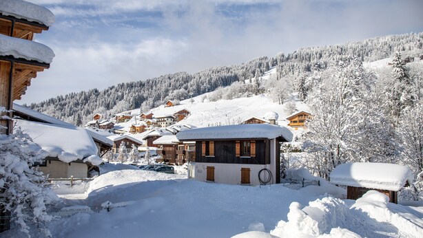 Ein verschneites Dorf mit Holzhäusern, einer kleinen Berghütte und einem Berg im Hintergrund.