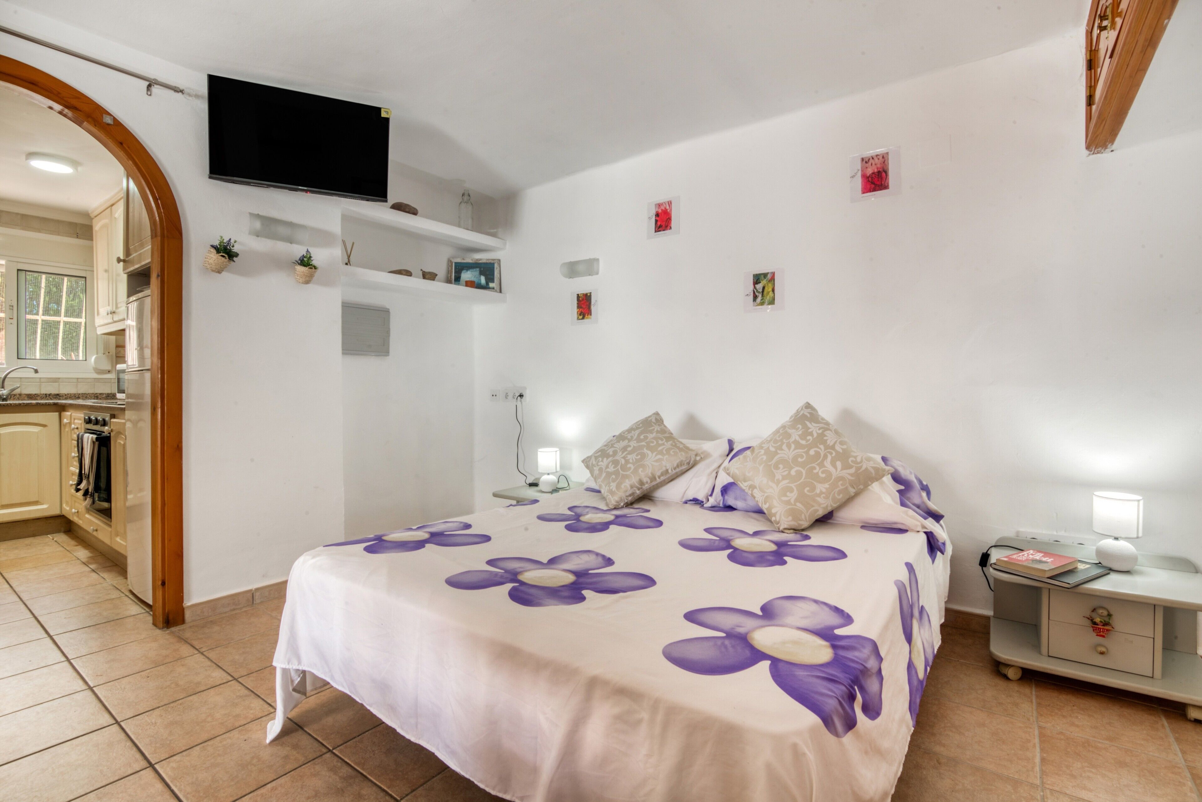 A bedroom with a bed, bedside tables, a TV, and a kitchen area visible through an archway.