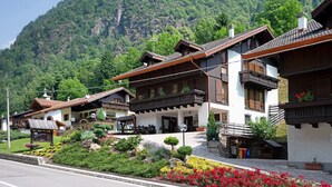 A traditional chalet-style hotel with a mountain backdrop, featuring balconies, a well-maintained garden, and a clear road in front.