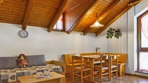 Een knus vertrek met een houten plafond, een bank, een tafel met stoelen, een klok en een raam met gordijn.