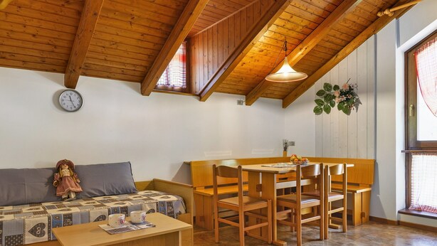 Ein gemütliches Zimmer mit Holzbalkendecke, einer Couch, einem Tisch mit Stühlen, einer Uhr und einem Fenster mit Vorhang.