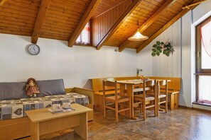 A cozy room with a wooden ceiling, a sofa, a table with chairs, a clock, and a window with a curtain.