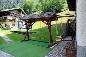 A wooden swing set with a roof, a stone bench, and a stone wall.