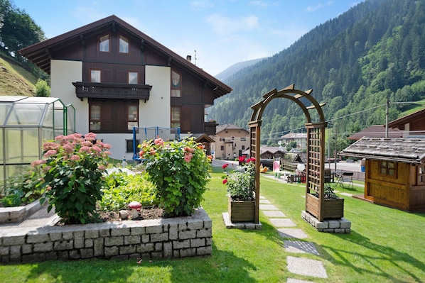 A chalet with a stone pathway leading to a wooden archway, surrounded by lush greenery and a mountain backdrop.