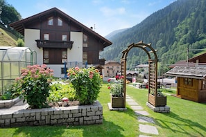 A chalet with a stone pathway leading to a wooden archway, surrounded by lush greenery and a mountain backdrop.