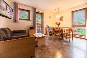 Een gezellige kamer met een bank, een eettafel met stoelen, een klein tafeltje met een tijdschrift, een stoel, een raam met gordijnen en uitzicht op groen buiten.