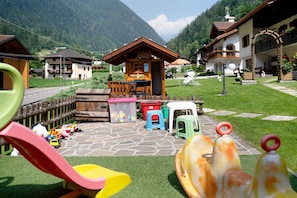 A playground with a slide and climbing structures, a wooden playhouse, and a stone pathway leading to a building with a balcony.