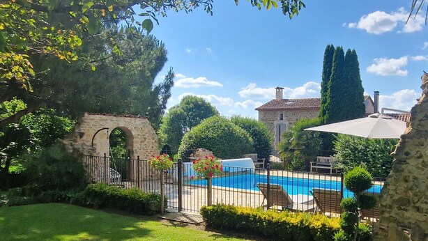 Una piscina con un arco de piedra, rodeada de follaje y un edificio de piedra al fondo.