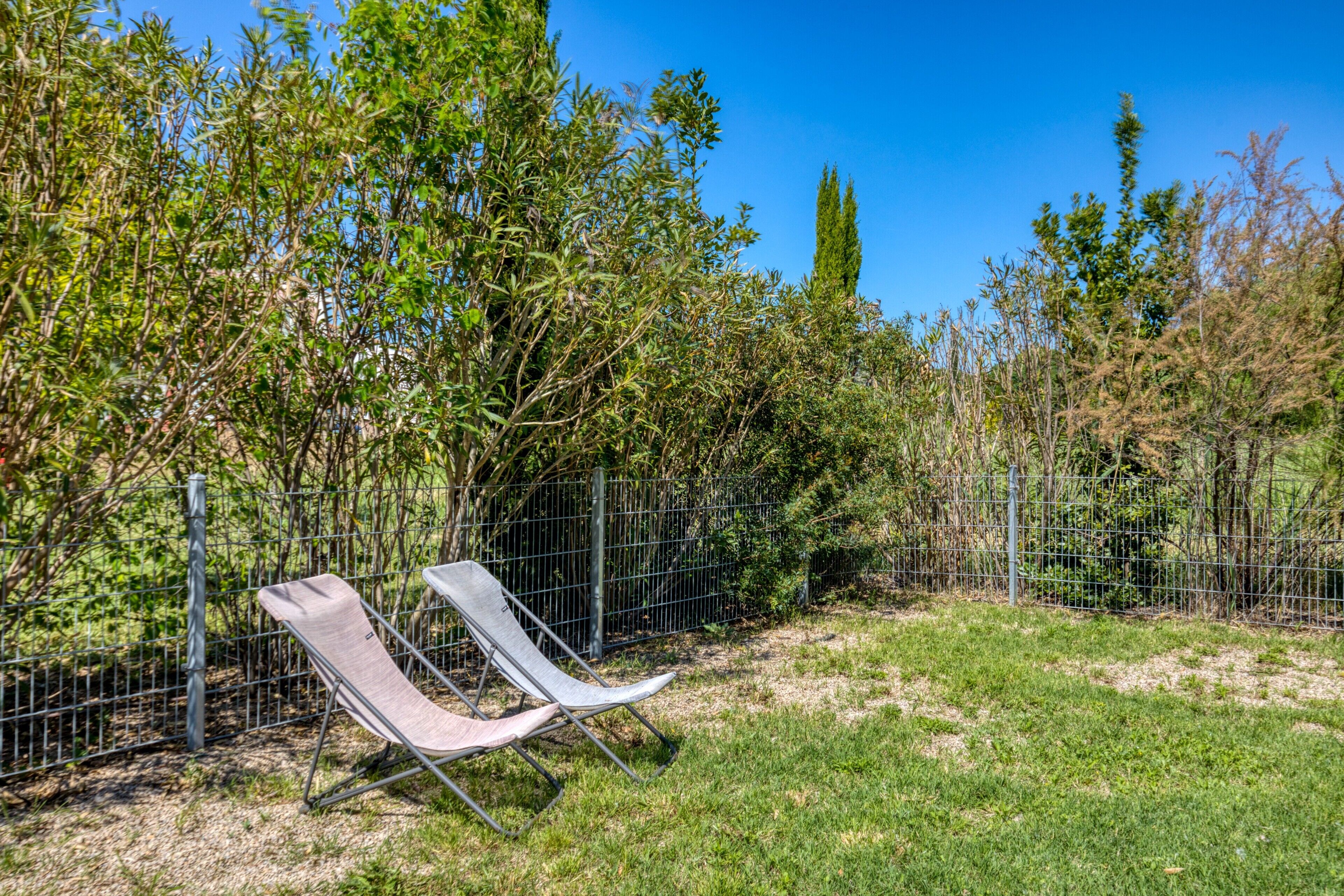 Twee metalen loungestoelen in een grasveld, met een metalen hek en bomen op de achtergrond.