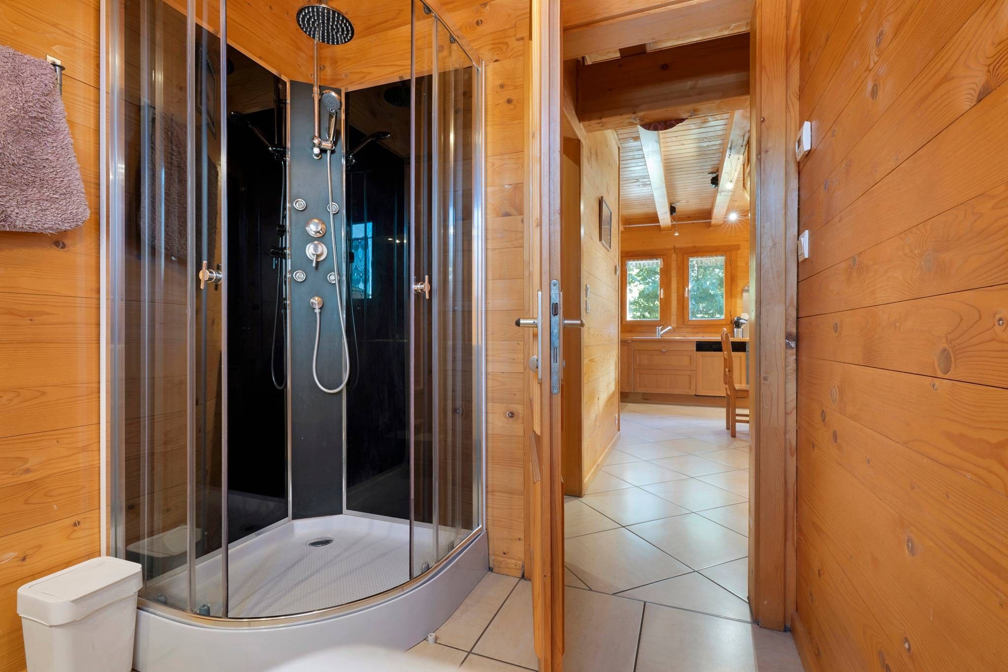 A wooden cabin interior with a glass-enclosed shower stall, a visible toilet, and a kitchen area with wooden cabinets and a window.