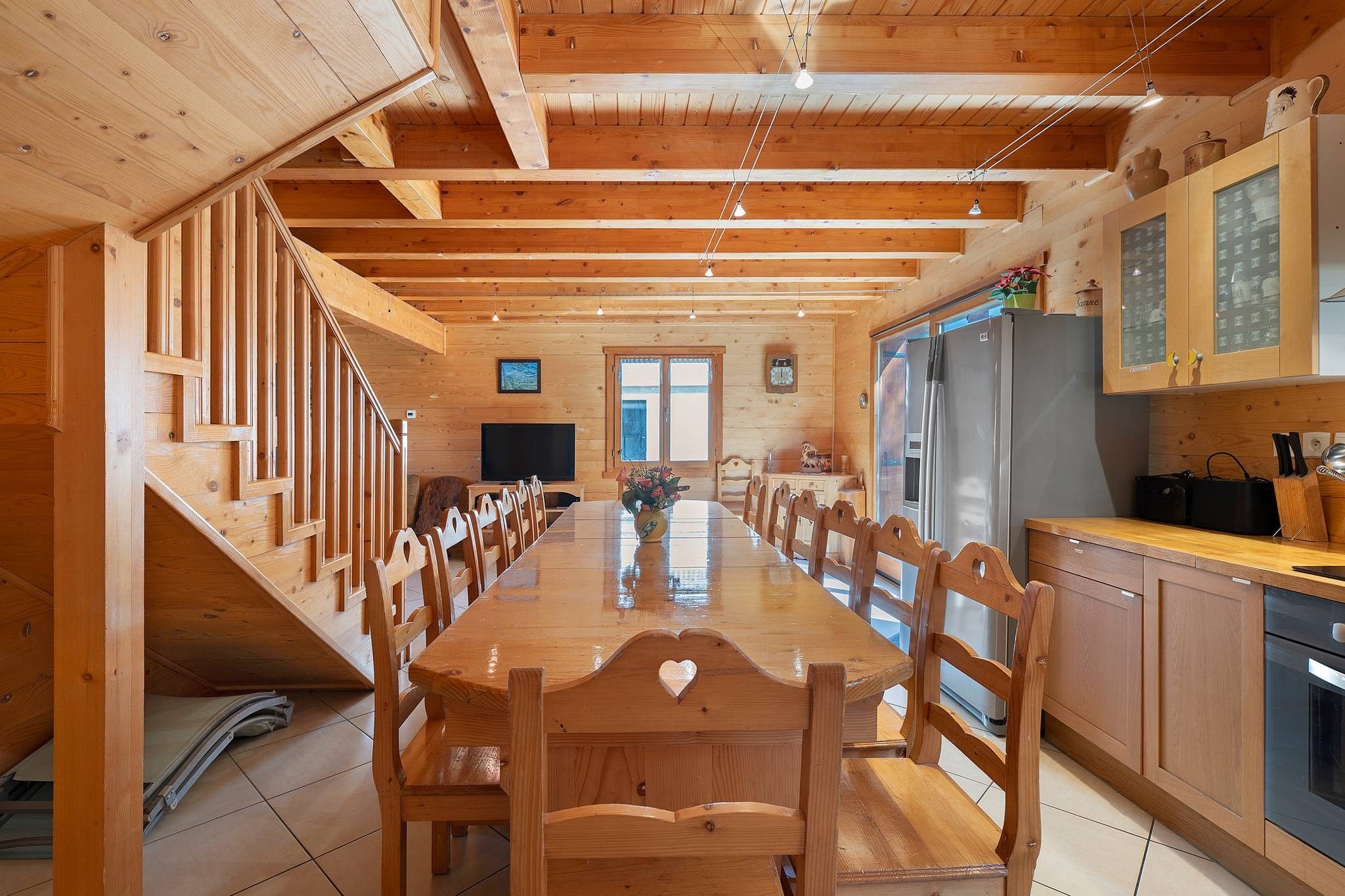A wooden interior with a long dining table, chairs, a kitchen area with cabinets, a refrigerator, and a television.