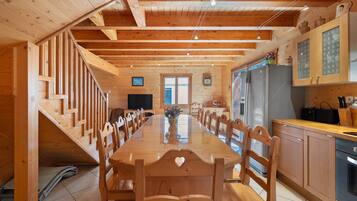 A wooden interior with a long dining table, chairs, a kitchen area with cabinets, a refrigerator, and a television.
