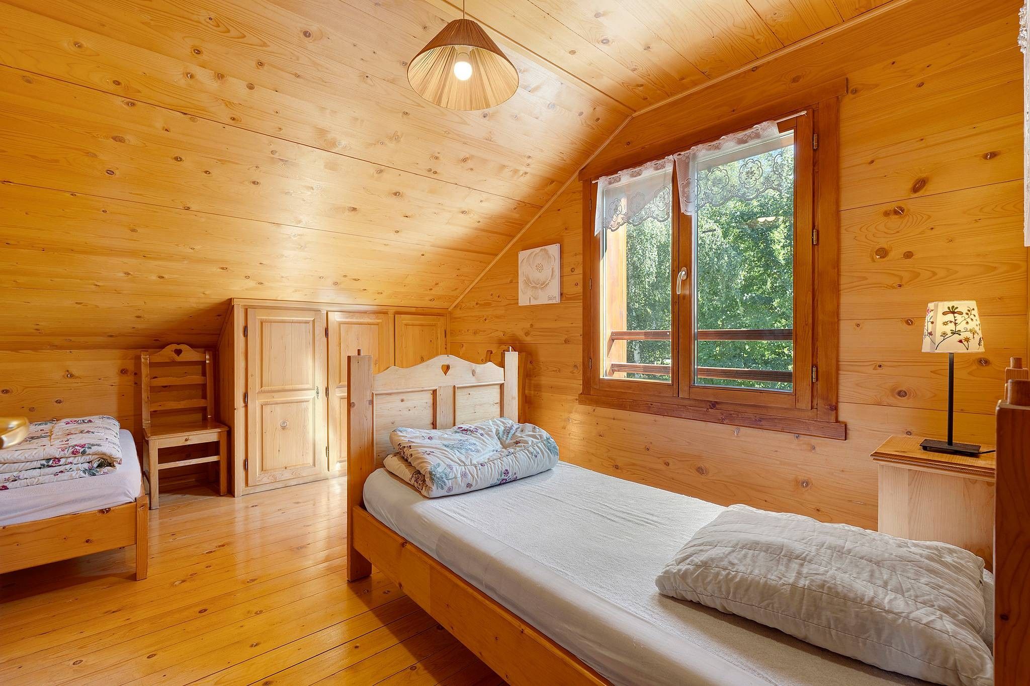 Acogedor dormitorio de cabaña de madera con una cama individual, cómoda, mesita de noche y ventana con vistas al follaje.
