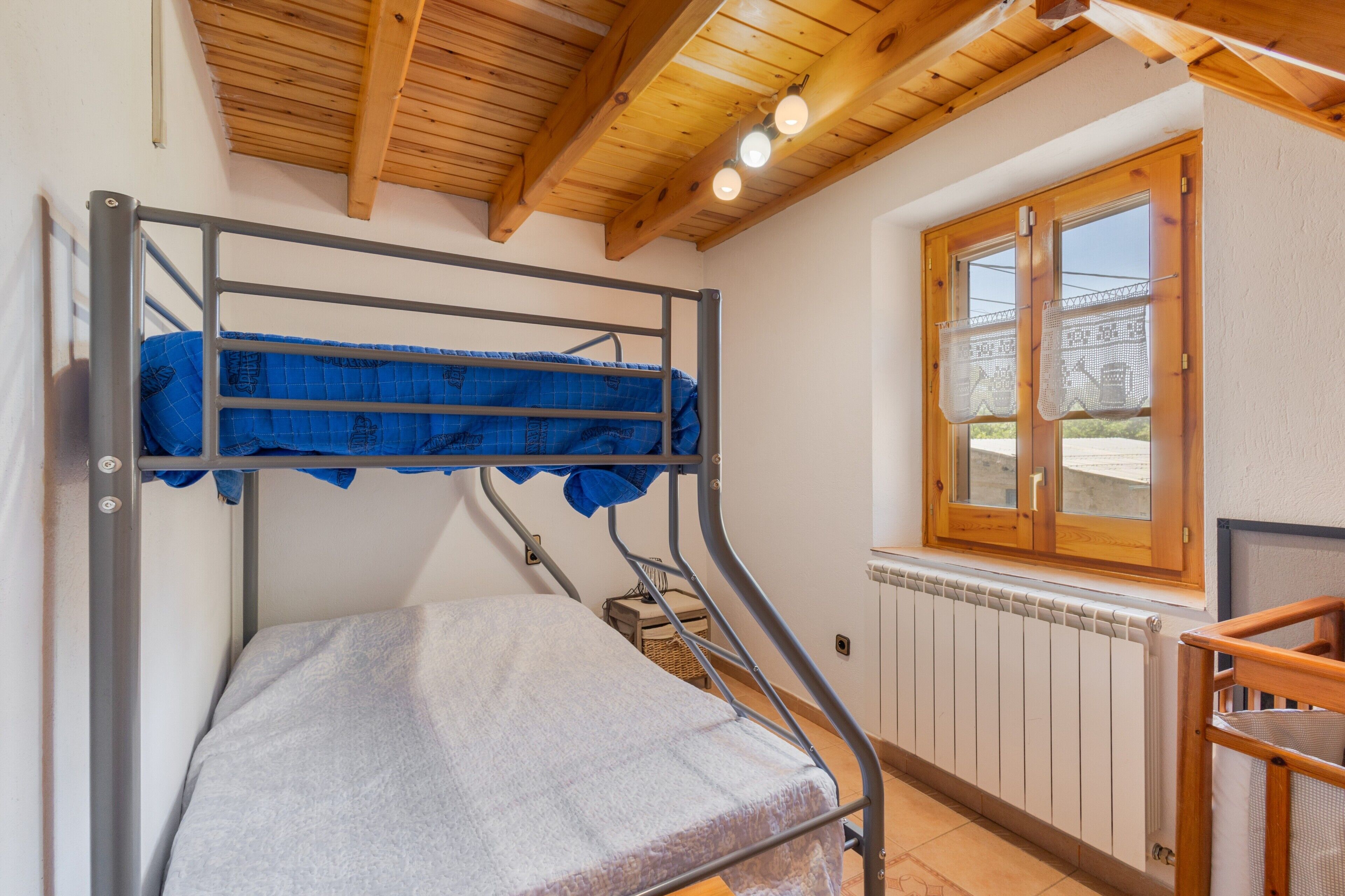 A bunk bed with a mattress, a wooden ceiling, a window with blinds, and a radiator.