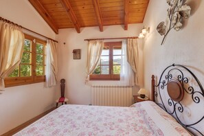 Et soveværelse med træloft, en seng med blomstret sengetæppe, en radiator og et vindue med gardiner.