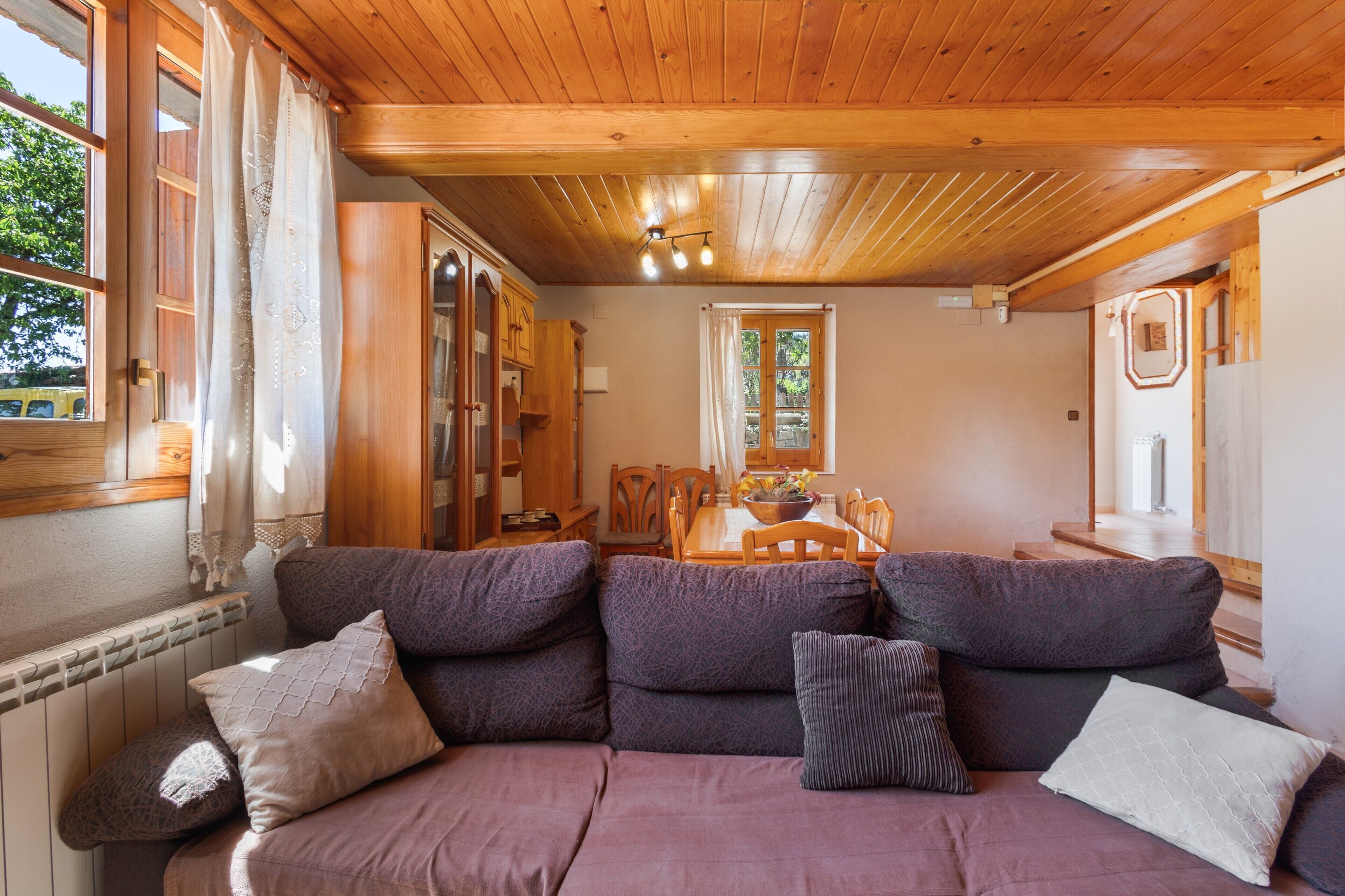 Een houten plafond, een bank met kussens, een eettafel met stoelen en een raam met gordijnen.