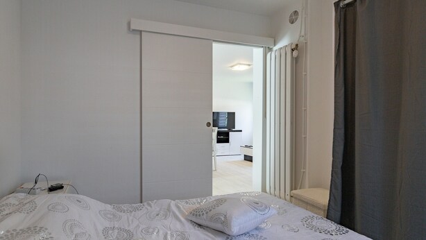 Une chambre à coucher avec un lit, une porte blanche, un téléviseur dans la pièce adjacente et une fenêtre avec des rideaux.