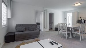 Un salon moderne avec un canapé gris, une table à manger avec des chaises et, au fond, un coin cuisine.