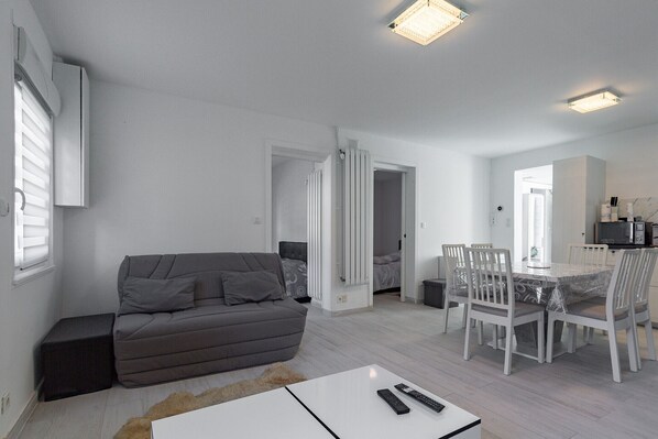 A modern living room with a grey sofa, a dining table with chairs, and a kitchen area in the background.