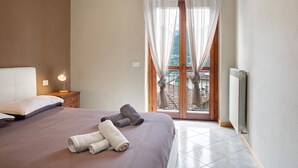A bedroom with a bed, a nightstand, a lamp, a window with sheer curtains, and a view of a balcony. - Apartment 'Typical  In Valtellina' with Mountain View, Private Terrace and Wi-Fi (Montagna In Valtellina)